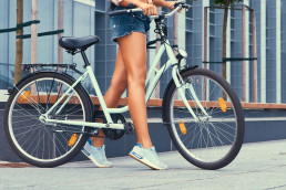 Cropped image of a slim girl with smooth long legs, wearing denim shorts, standing with city bike against a skyscraper
