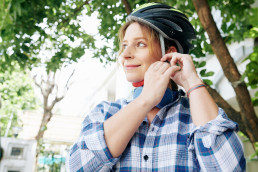 Smiling pretty young woman in plaid shirt putting on bicycle helmet
